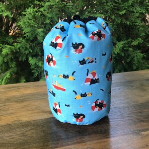 May include: A blue drawstring bag with a pattern of black cats wearing life preservers and swimming in the ocean. The bag is made of fabric and has a drawstring closure.
