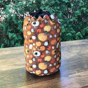 May include: A brown drawstring bag with a black drawstring. The bag is covered in a pattern of orange, yellow, and white pumpkins and black cats.