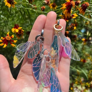 May include: A pair of iridescent butterfly wing earrings with silver and gold hoops. The wings are translucent and have a rainbow sheen.