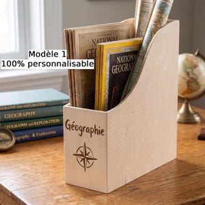 May include: A light-colored wooden magazine holder featuring the engraved word Géographie and a compass rose symbol on the front. The holder contains several National Geographic magazines and is positioned next to a stack of books and a compass.