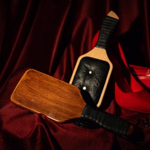 Puede incluir: Dos paletas de madera con mangos y detalles de cuero negro, una con una superficie de golpeo de cuero negro, descansan sobre una superficie de terciopelo rojo oscuro. Un par de zapatos de tacón rojos son visibles en el fondo.