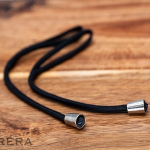Può includere: Un cordino nero con estremità metalliche argentate, appoggiato su una superficie di legno. Il cordino è realizzato in materiale intrecciato e sembra flessibile. Le estremità sono cilindriche e lisce.