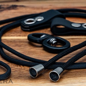 May include: Black lanyard with metal accents and leather attachments. The lanyard features a braided cord design and silver-colored metal tips. The leather attachments have silver-colored rivets. The items are arranged on a wooden surface.