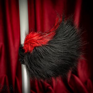 Peut inclure: Accessoire en fausse fourrure noire et rouge, peut-être un élément de costume, sur fond rouge foncé. L'accessoire a une bande de fourrure rouge vif sur une plus grande section de fourrure noire. Le mot "VARERA" est visible en bas à gauche.