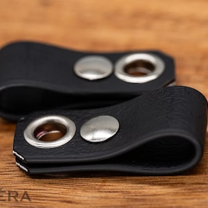 Op de afbeelding: Twee zwarte leren kabelorganizers met zilveren metalen oogjes en druksluitingen. De organizers liggen op een houten oppervlak. Het leer heeft een textuur.