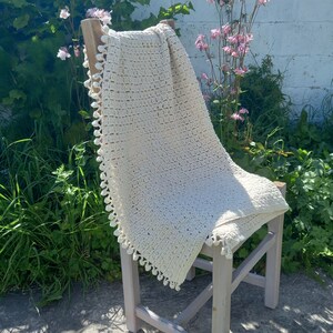 May include: A white crocheted throw blanket with a fringe border draped over a wooden chair. The blanket has a delicate, lacy pattern and is perfect for adding a touch of warmth and texture to any room.