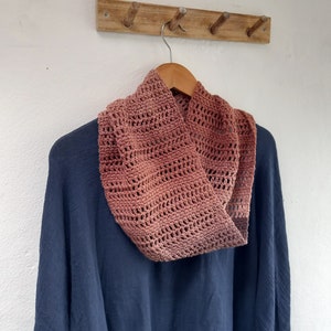 Peut inclure: Un col en crochet de couleur terre cuite, suspendu à un cintre en bois sur un mur blanc.