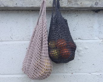 May include: Two crocheted string bags hanging on a wooden surface. The bag on the left is pink and the bag on the right is gray. The gray bag is filled with red and orange produce.