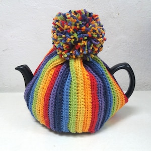 Puede incluir: Un calienta-tetera de punto de crochet a rayas de arcoíris con un pompón en la parte superior.