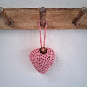 May include: A handmade, pink crocheted heart ornament with a wooden button detail. The heart is suspended by a matching pink cord and hangs from a wooden peg rack. The heart is approximately 3 inches wide.