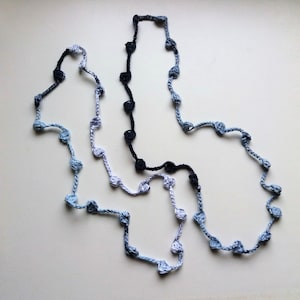May include: A crocheted necklace with three strands of different colors: black, light blue, and dark blue. The necklace is in the shape of a figure eight.