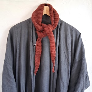May include: A dark charcoal gray open-front jacket with long sleeves and a relaxed fit, layered with a rust-colored crocheted scarf tied in a simple knot. The scarf features a textured, repeating stitch pattern and tapered ends, draped over a wooden hanger.