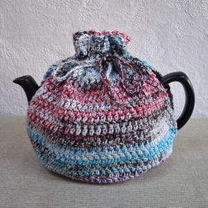 May include: A multi-colored, crocheted tea cozy in shades of red, blue, gray, and white, covers a black teapot. The cozy has a drawstring closure at the top and a striped pattern. The teapot's handle and spout are visible.