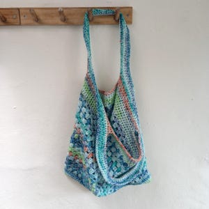 May include: A handmade crochet tote bag in shades of blue, green, and coral hangs from a wooden peg rack. The bag features a textured stitch pattern and long straps for carrying over the shoulder. The bag is a unique, eco-friendly accessory.