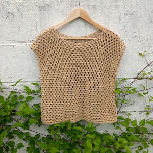 Puede incluir: Un top de crochet beige con un tejido suelto y abierto. El top tiene un escote redondo y es sin mangas. Está colgado en una percha de madera contra una pared blanca.
