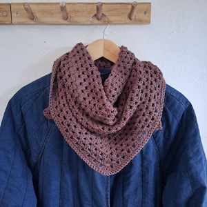 May include: A dusty rose crochet cowl scarf with an open, airy design. The scarf is triangular and hangs on a wooden hanger. The background includes a blue jacket and a wooden wall-mounted rack with pegs.