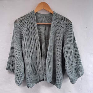 May include: A sage green open-front cardigan featuring a textured crochet stitch pattern. This garment has three-quarter length sleeves and a relaxed, loose-fitting silhouette. The piece is displayed on a wooden hanger against a plain background.