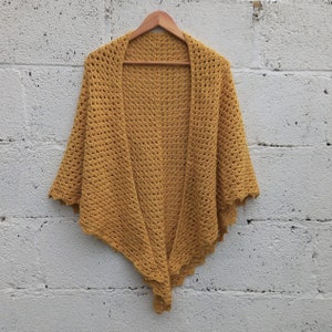 May include: A mustard yellow crochet shawl with a scalloped edge. The shawl is hanging on a wooden hanger against a white brick wall.