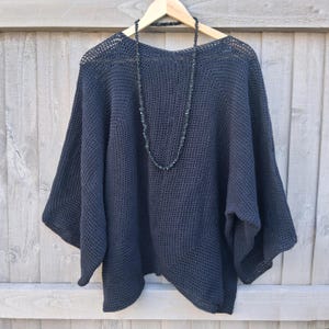 May include: A dark charcoal gray crochet top featuring a wide, loose-fitting silhouette with three-quarter length sleeves and a wide neckline. A long, thin necklace made of small, dark, irregular-shaped beads hangs over the front of the garment.