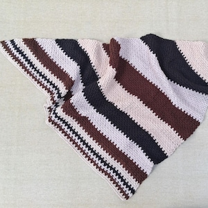 May include: A hand-crocheted blanket featuring a striped design in brown, white, and navy blue. The blanket is folded, displaying the diagonal stripes and textured stitches. The colour scheme is neutral and earthy.
