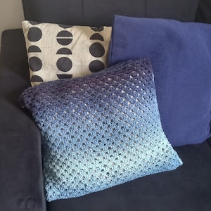 May include: A blue and teal crocheted cushion with a square pattern. The cushion is on a dark blue sofa with two other cushions behind it.