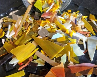 May include: A collection of assorted stained glass shards in shades of yellow, amber, orange, and white, spilling out from a clear glass bowl. A single silver-colored coin is placed among the scattered, irregularly shaped glass pieces.
