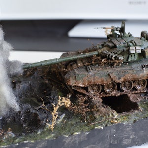 Op de afbeelding: Een model van een groene tank met een camouflagepatroon, bedekt met modder, met een witte rookpluim erachter. De tank staat op een dioramabasis met bruine aarde en groen gebladerte.