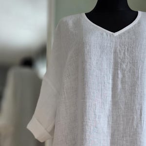 May include: A white linen tunic with a V-neck and rolled-up sleeves. The lightweight fabric drapes loosely, suggesting a relaxed fit. The garment is displayed on a black mannequin, highlighting the crisp, clean lines of the design.