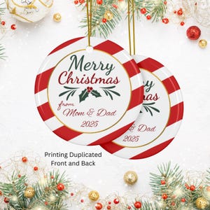 Puede incluir: Dos adornos navideños redondos con rayas rojas y blancas. Los adornos tienen un borde dorado y el texto "Merry Christmas from Mom & Dad 2025". Los adornos cuelgan de cordones dorados.