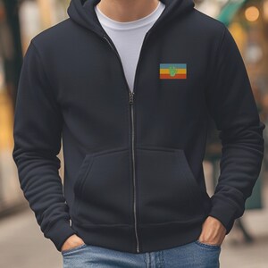 May include: A black zip-up hoodie with a small embroidered patch on the left chest. The patch features a yellow, red, and blue striped background with a green handprint in the center.
