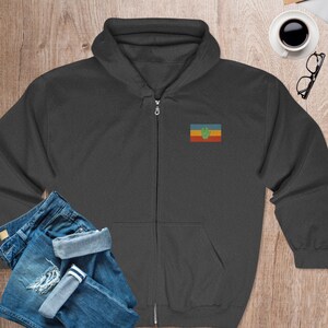 May include: A dark gray zippered hoodie with a small embroidered flag on the left chest. The flag is a horizontal tri-color of yellow, blue, and red with a green handprint in the center.