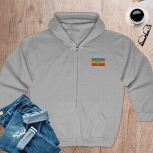 May include: A gray zippered hoodie with a small embroidered flag on the left chest. The flag is red, yellow, green, and blue with a green handprint in the center.