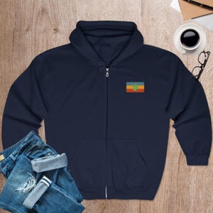 May include: A navy blue zip-up hoodie with a small embroidered flag on the left chest. The flag has a green handprint on a yellow, red, and blue background.