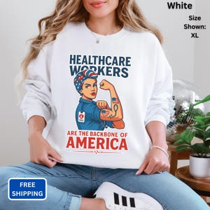 May include: White sweatshirt with a graphic of a healthcare worker flexing their arm, with the text "HEALTHCARE WORKERS ARE THE BACKBONE OF AMERICA." The design features red, white, and blue colors, and a medical symbol.