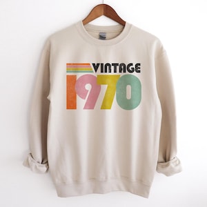 May include: A beige sweatshirt with a vintage 1970 graphic in rainbow colors. The text "Vintage" is in black.