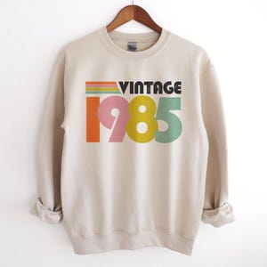 May include: A beige sweatshirt with a vintage 1985 graphic in rainbow colors. The text "Vintage" is in black.