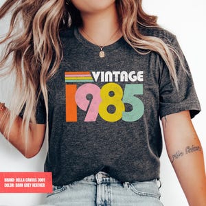 May include: Dark grey heather t-shirt with the word "VINTAGE" above the colorful number "1985". The shirt has a retro design with a rainbow stripe above the numbers. The shirt is made of Bella Canvas 3001 material.