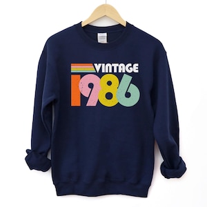 May include: Navy blue sweatshirt featuring the word "VINTAGE" in white and the year "1986" in colourful block letters. The sweatshirt is hanging on a wooden hanger against a white background.