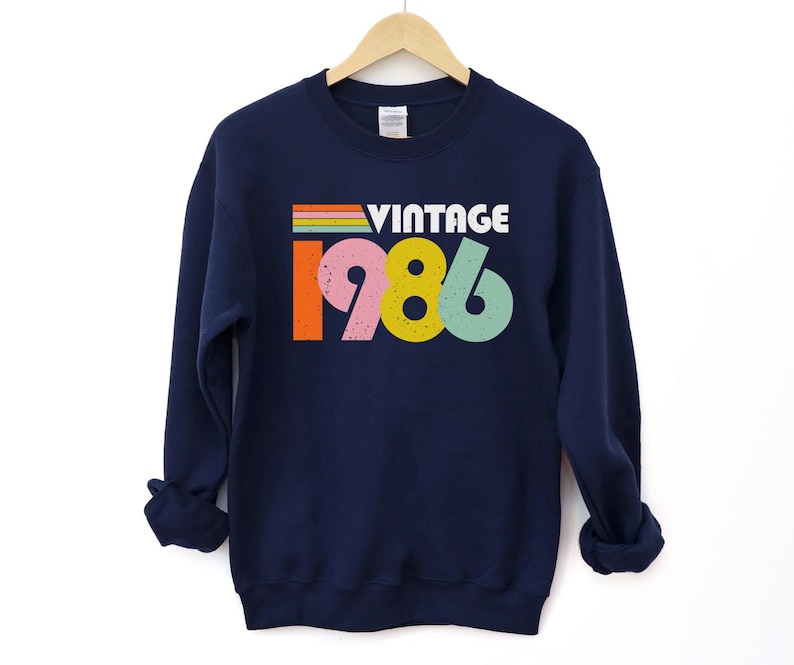 May include: Navy blue sweatshirt featuring the word "VINTAGE" in white and the year "1986" in colourful block letters. The sweatshirt is hanging on a wooden hanger against a white background.