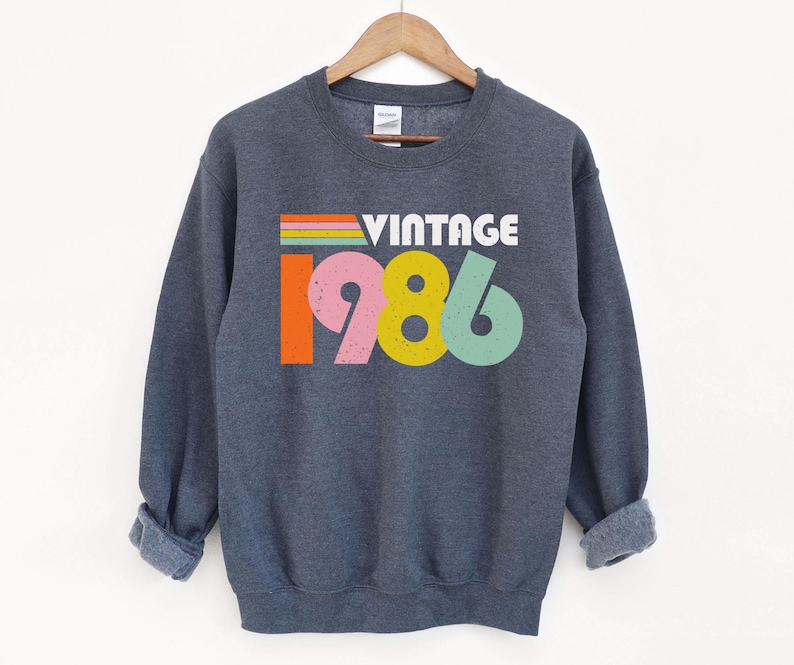 May include: A dark grey sweatshirt featuring the word "VINTAGE" above the number "1986" in a colourful design. The sweatshirt has long sleeves and a crew neck, and is suspended from a wooden hanger.