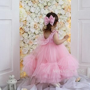 May include: A young girl wearing a pink tulle dress with a sparkly bodice and a large pink bow in her hair. She is standing in front of a wall of white and peach roses.