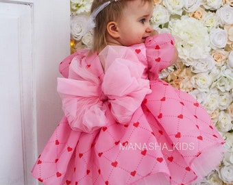 Puede incluir: Vestido rosa de bebé con estampado de corazones y un gran lazo en la espalda. El vestido tiene una falda de tul y se lleva con una diadema a juego. El texto "MANASHA_KIDS" es visible en el vestido.