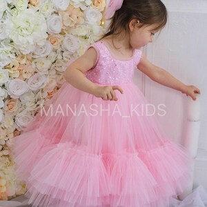 May include: A little girl wearing a pink tulle dress with a sequined bodice. The dress has multiple layers of tulle and is very full. The girl is standing in front of a white flower wall.