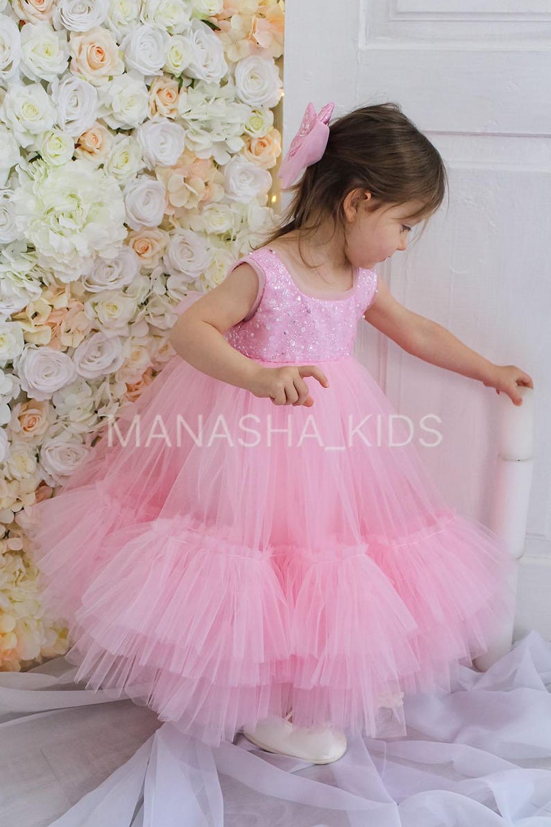 May include: A little girl wearing a pink tulle dress with a sequined bodice. The dress has multiple layers of tulle and is very full. The girl is standing in front of a white flower wall.