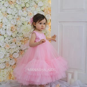 May include: A young girl wearing a pink tulle dress with sequins. The dress has multiple layers of tulle and is very full. The girl is standing in front of a white flower wall and a white door. The text "MANASHA_KIDS" is visible on the dress.