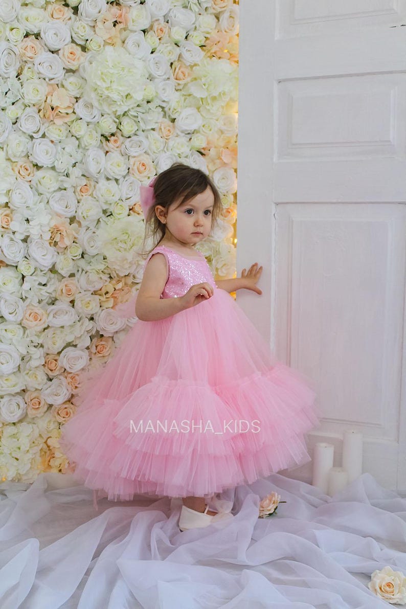 May include: A young girl wearing a pink tulle dress with sequins. The dress has multiple layers of tulle and is very full. The girl is standing in front of a white flower wall and a white door. The text "MANASHA_KIDS" is visible on the dress.
