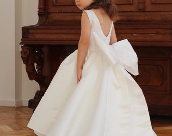 May include: A white satin flower girl dress with a large bow on the back. The dress has a V-neck and a flowing skirt. The dress is worn by a young girl in front of a wooden piano. The text "MANASHA_KIDS" is visible.