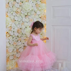 May include: A young girl wearing a pink tulle dress with sequins on the bodice. The dress has multiple layers of tulle and is very full. The girl is standing in front of a white flower wall with the text "MANASHA_KIDS" visible in the bottom right corner.