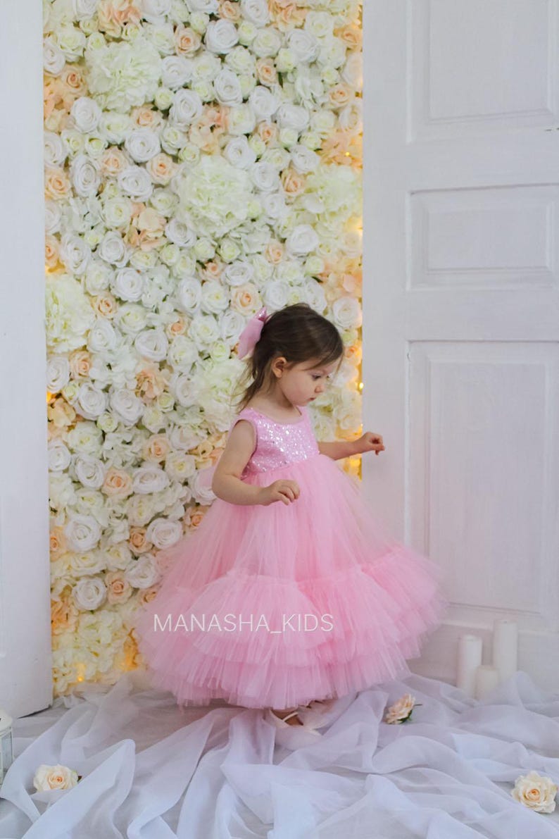 May include: A young girl wearing a pink tulle dress with sequins on the bodice. The dress has multiple layers of tulle and is very full. The girl is standing in front of a white flower wall with the text "MANASHA_KIDS" visible in the bottom right corner.