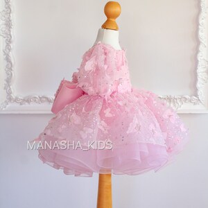 May include: A pink tulle dress with butterfly embellishments and a large pink bow. The dress is on a wooden mannequin.